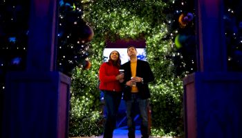 A couple stands on a glowing, festive stairway lined with twinkling lights and trees, smiling and holding drinks in a celebratory moment.