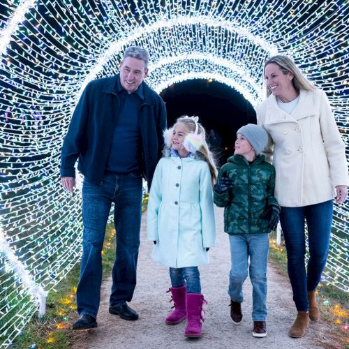 A family of four walks through a tunnel of twinkling lights, smiling and holding hands as they explore a festive, illuminated walkway.