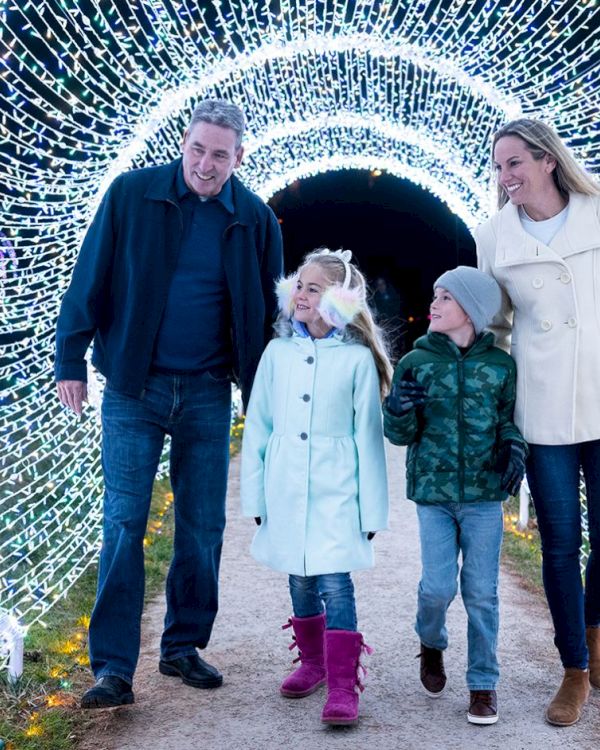 A family of four walks through a tunnel of twinkling lights, smiling and holding hands as they explore a festive, illuminated walkway.