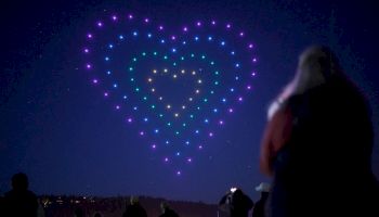 A night sky displays a heart-shaped formation of colorful lights, with people watching and silhouettes in the foreground.