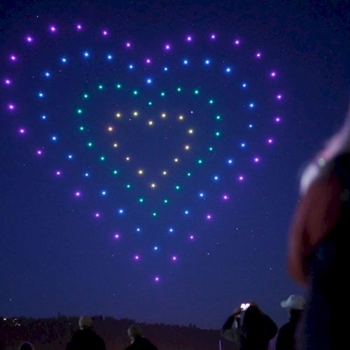 A night sky display with layered heart-shaped light patterns in purple and blue, created by numerous bright points; silhouettes of spectators below.