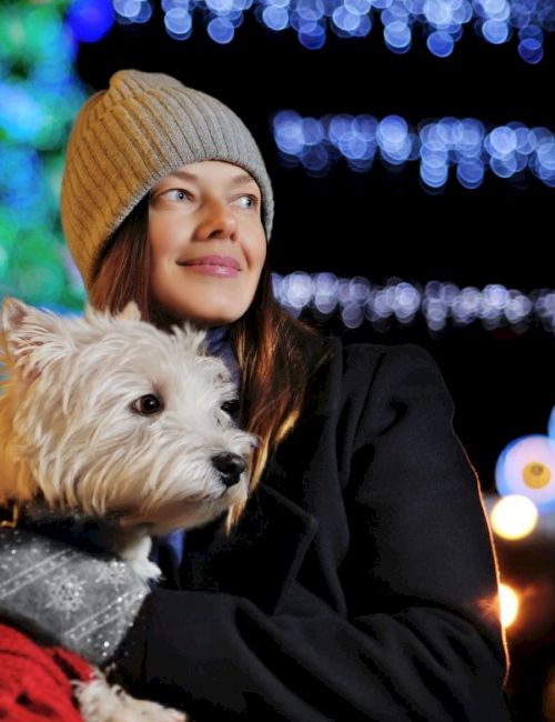 A woman in a beanie holds a small dog wrapped in a red blanket, at a festive outdoor night scene with lights and a Christmas tree.