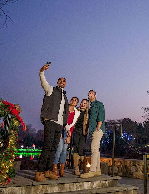A group of friends takes a selfie beside a tall, illuminated Christmas tree in a festive outdoor setting, with a decorated archway nearby.