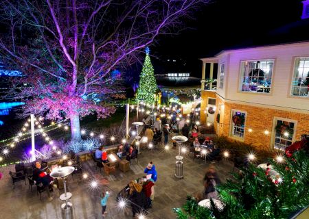 A night outdoor cafe scene with string lights, diners at tables, a lit Christmas tree, festive trees, and a brick building patio crowding around festive lights.
