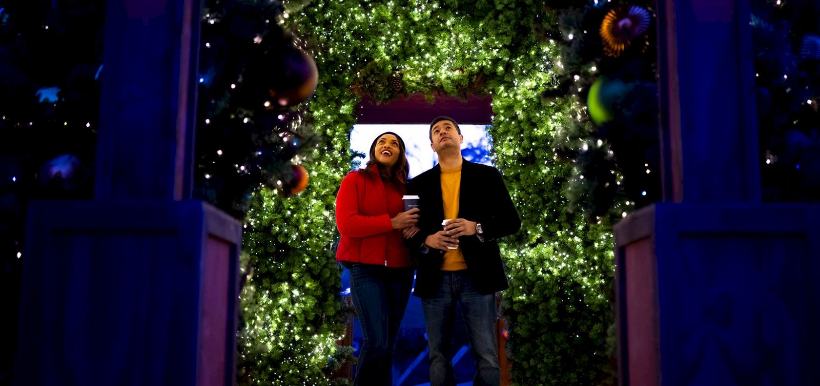 A couple stands under a glowing, tree-lit archway with festive lights and hanging ornaments, holding drinks and smiling at the camera.
