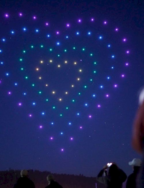 A dark night shows a large heart-shaped display of purple and blue lights in the sky, with people watching and a woman in the foreground.