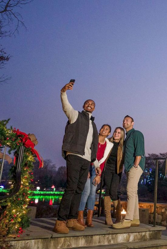 A group of four people taking a selfie near a tall, decorated Christmas tree at an outdoor holiday park, with festive lights and a wooden entrance.