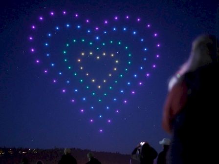 A night scene shows a person in foreground watching a large heart-shaped display of colorful lights in the sky.