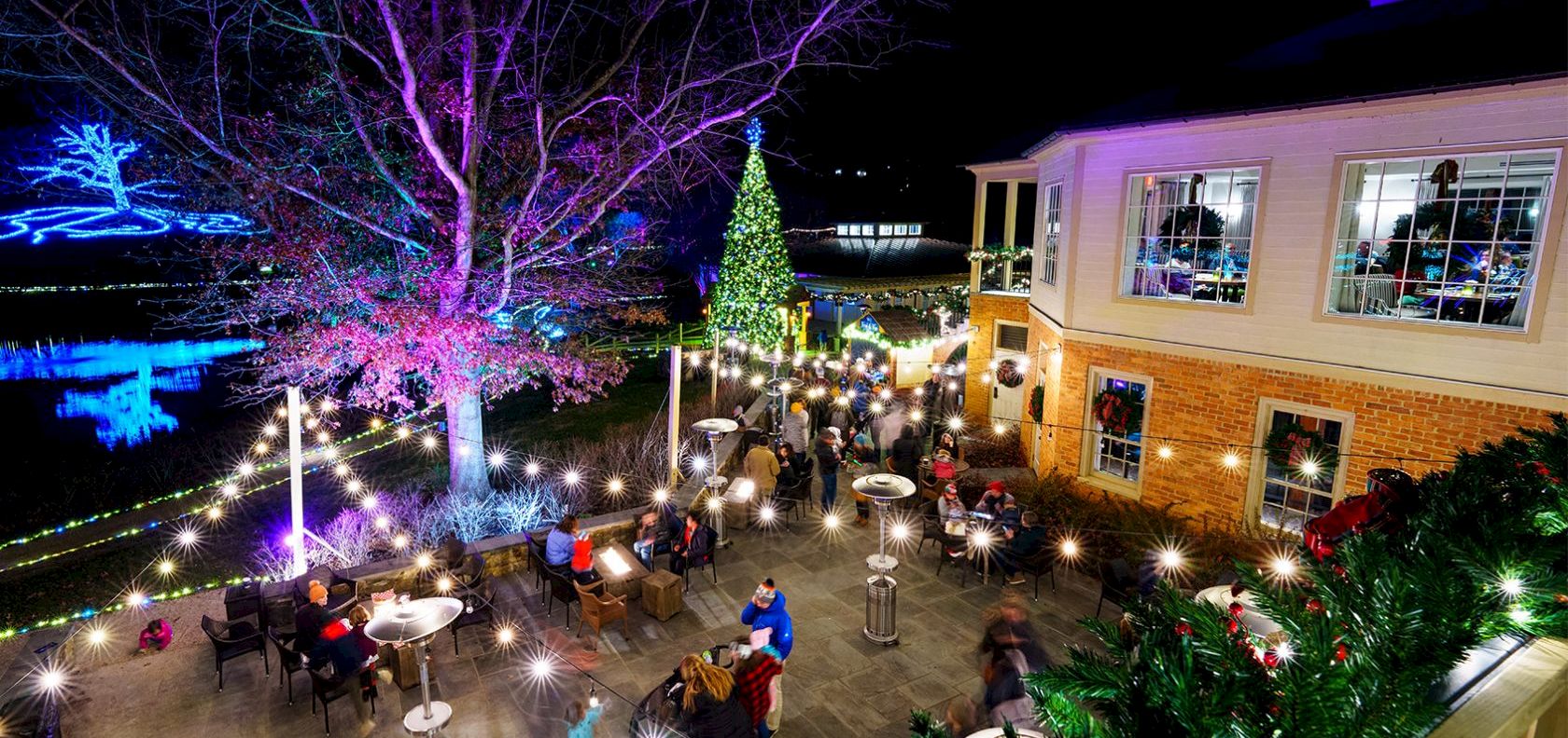 People dining and mingling at an outdoor nighttime venue, string lights glowing, a tall Christmas tree, festive decor, and a brick building lit up in the background.