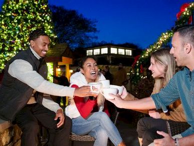 A group of four friends sharing drinks and laughs around a small table outdoors at night, with festive lights and trees in the background.
