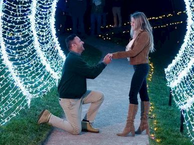 A man proposes on a lit tunnel of fairy lights, kneeling on one knee while a woman stands, smiling, in a festive outdoor setting.