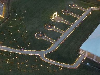 An aerial view of a festive event venue: glowing pathways, decorative lights, a large white tent, a blue-lit entrance, and landscaped grounds.