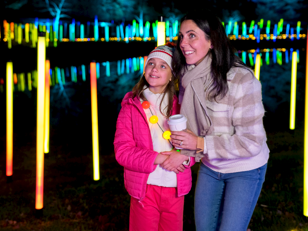 Two people, a woman and a girl, stand under glowing colorful light poles, smiling and posing together at night.