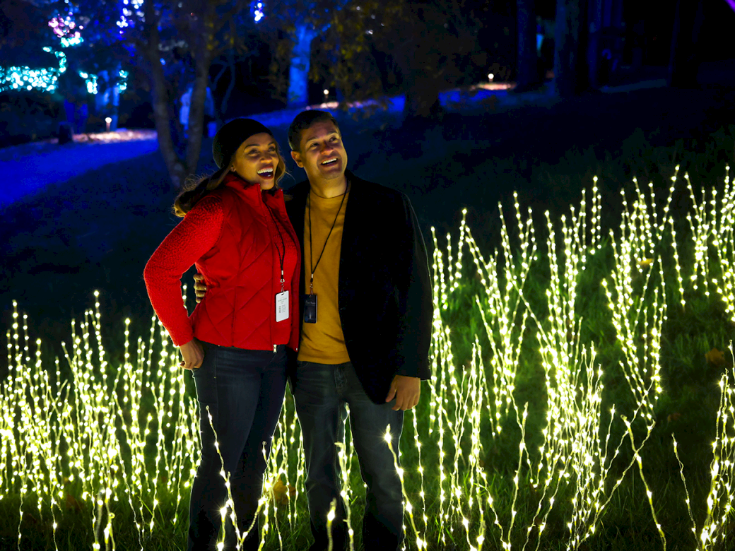 Two people posing happily among bright, twinkling lights outdoors at night, smiling for the camera as they stand close together.