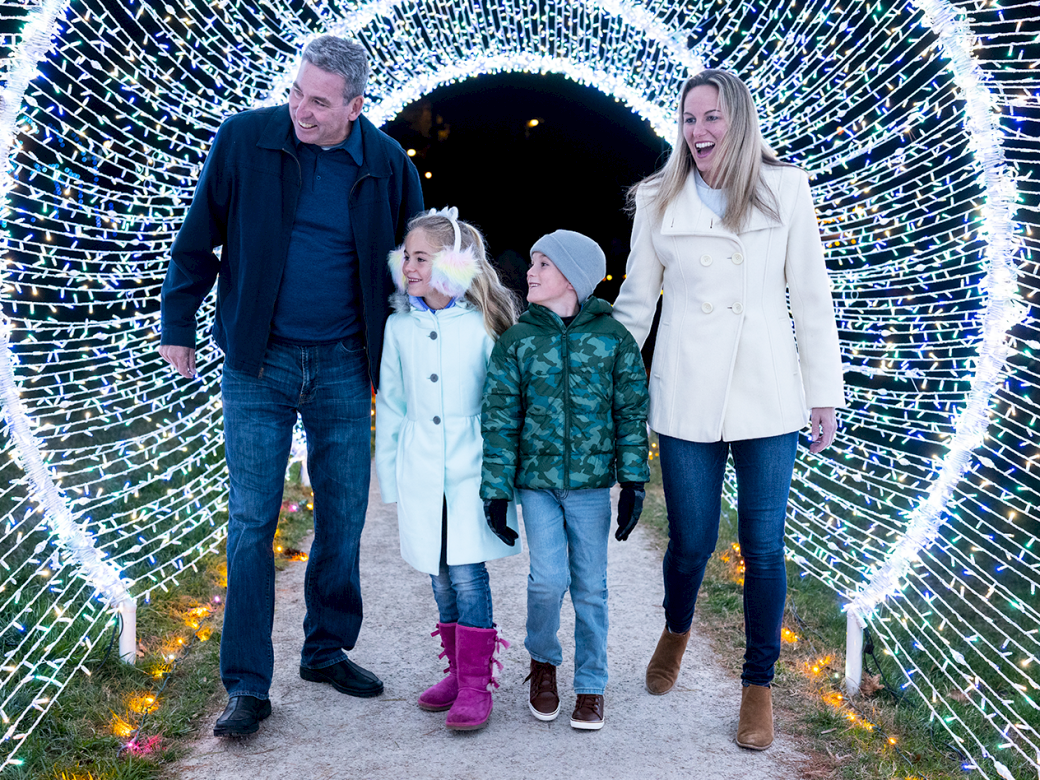 A family of four walks through a glowing tunnel of lights outdoors, smiling and holding hands, wearing coats and boots.