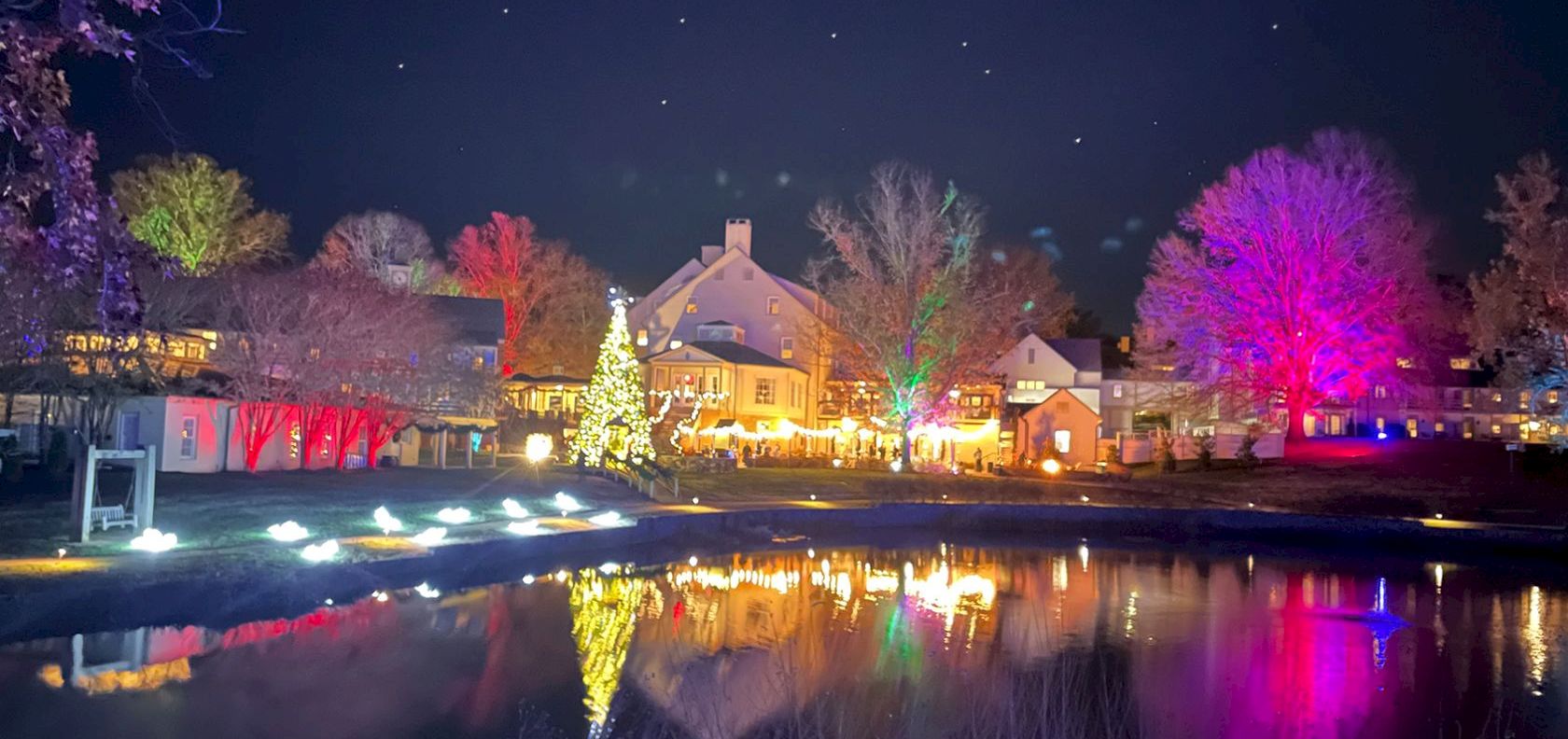 A festive village by a calm pond, with colorful lights, a glowing Christmas tree, and a starry night sky.