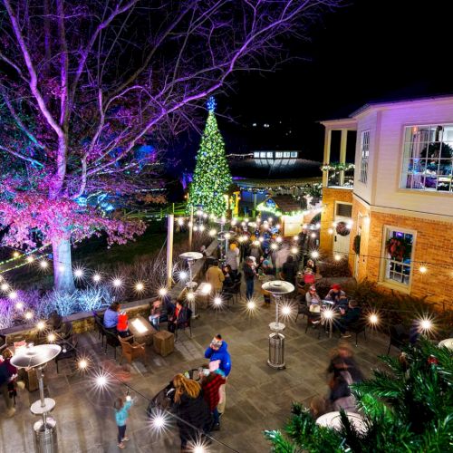 A festive outdoor cafe at night with string lights, a lit Christmas tree, people dining, and a brick building, festive decor and trees glow.