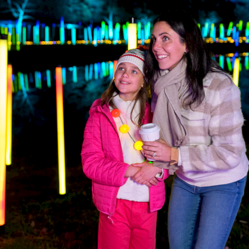 Two people, a woman and a child, stand among glowing, colorful light tubes at night, smiling and posing for a photo.