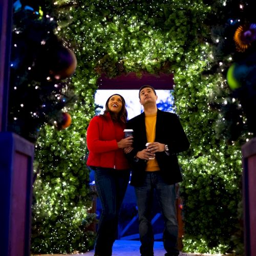A couple stands under a sparkling arch of Christmas lights, smiling with drinks in hand, immersed in a festive, twinkling glow.
