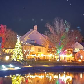 A festive village at night with colorful lights on trees and a central church, reflecting off a calm pond.