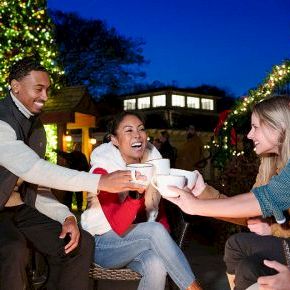 Four friends share a joyful outdoor evening, toasting cups under festive lights and trees.
