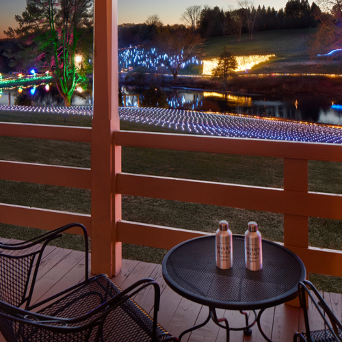 A balcony table and chairs overlook a lake or park with colorful holiday lights reflecting on the water at dusk.