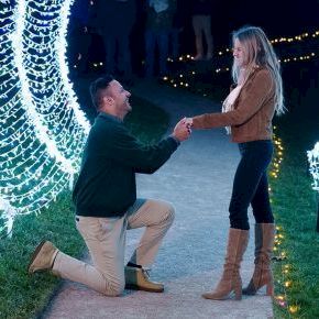 A man proposes on a lit tunnel path, kneeling with a ring as a woman stands surprised under bright string lights.