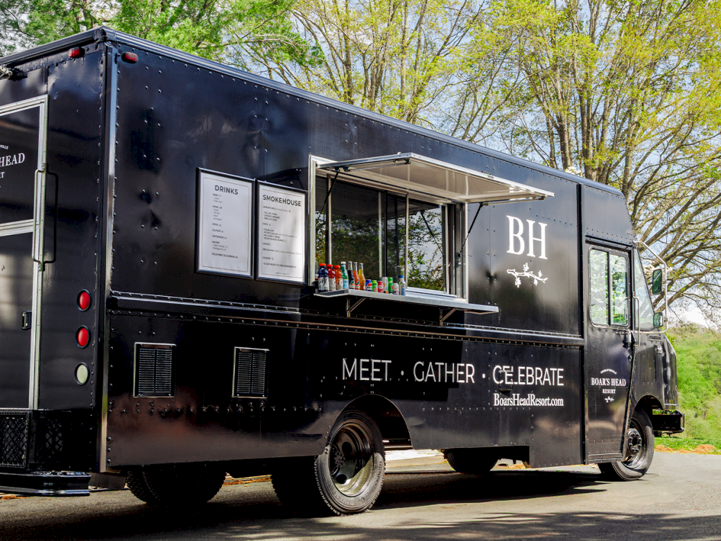 A black food truck with a window service counter, listing &ldquo;MEET &bull; GATHER &bull; CELEBRATE&rdquo; and &ldquo;BH&rdquo; on the side, parked by trees.