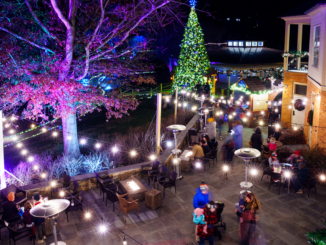 Outdoor nighttime cafe or restaurant patio with string lights, a decorated Christmas tree, and people socializing near festive tables.