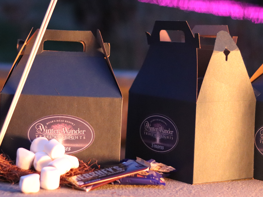 A row of dark gift boxes with handles, each labeled, plus hot chocolate mix, marshmallows, and candy, all set on a surface at dusk.